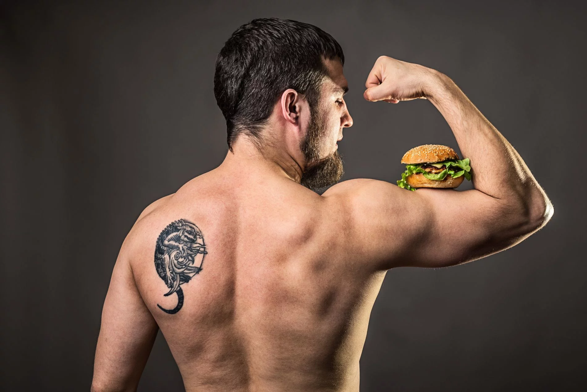 Izmos, szakállas férfi hátulnézetből, megfeszített bicepszén hamburgerrel – edzés és táplálkozás