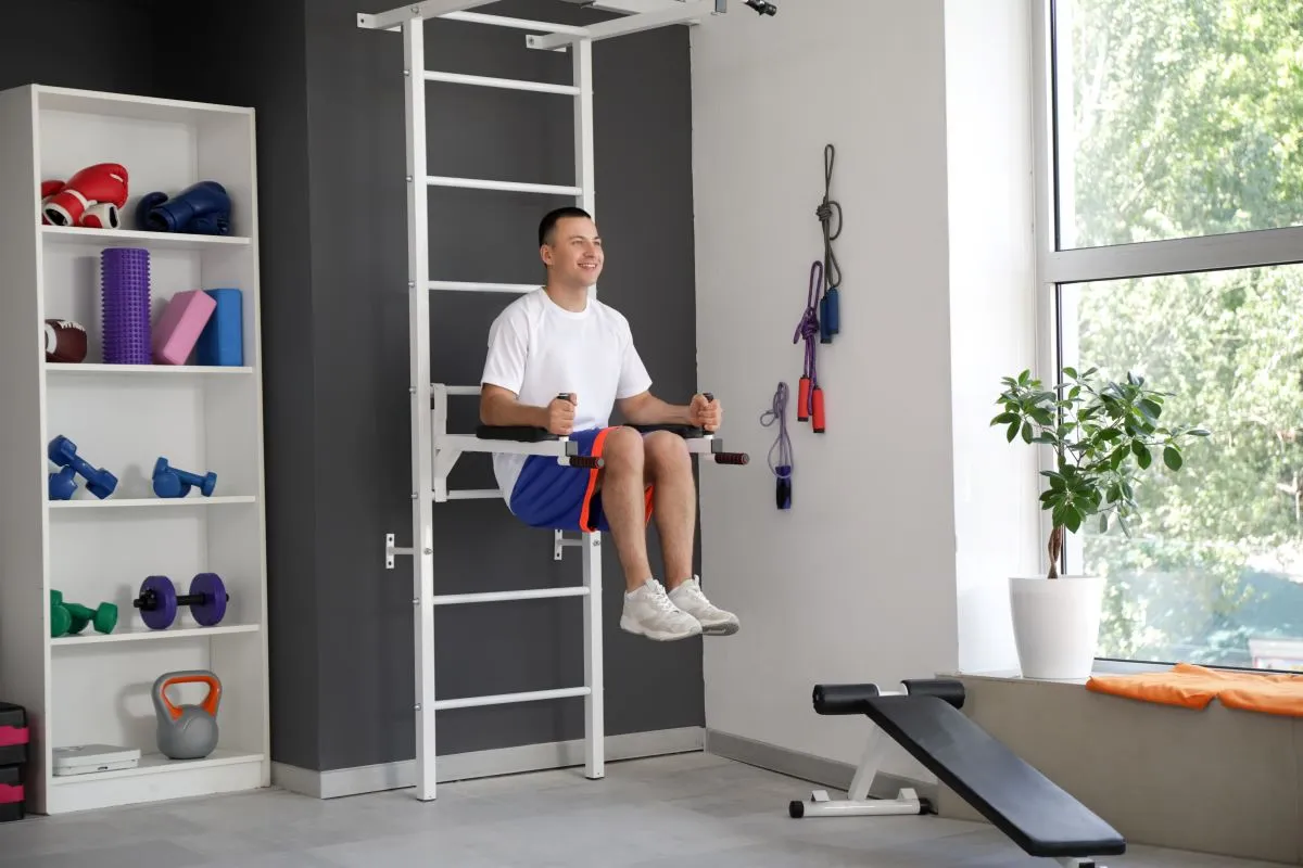 Férfi lábemelést végez a bordásfalra szerelt tolódzkodón egy otthoni edzőszobában, a polcon kettlebell, súlyzók és tornakellékek